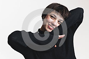 Dreamy woman with short brown hair in basic black turtleneck smiling and touching her face with hand isolated over white