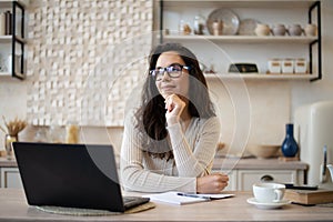 Dreamy woman sitting in kitchen at table with opened laptop, looking away and thinking, copy space