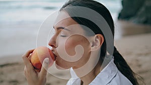 Dreamy woman tasting peach sitting on sand beach at summer vacation closeup