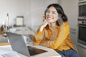 Dreamy Young Arab Female Sitting At Desk With Laptop In Kitchen