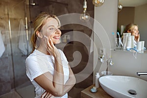 Dreamy young beautiful blonde woman smiling at mirror in bathroom. Self care concept.