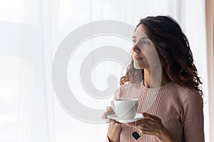 Dreamy young beautiful woman standing near window with cup of tea.