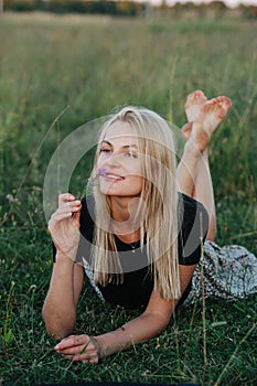 Dreamy young blond woman lying on a grass in a field, smelling wild flower