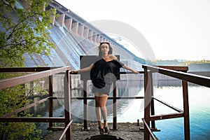 A dreamy young brunette girl stay near hydroelectric power station. Evening time. Side view