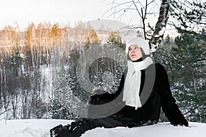 Dreamy young girl is siting on snow in winter sunny evening forest and is looking up.