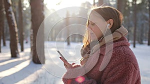 Dreamy young woman with frosty breath is using smartphone and listening to music is using headphones in winter park