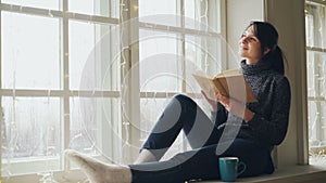Dreamy young woman is reading book and smiling on Christmas sitting on window-sill decorated with illumination. Girl is