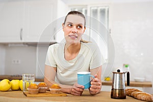 Dreamy young woman standing in home kitchen with cup of coffee