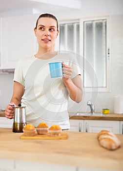 Dreamy young woman standing in home kitchen with cup of coffee