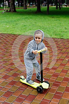 Drenched in the rain, a boy in a sport suit skates on a scooter. Spring walk in the city park, rainy weather.