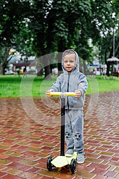 Drenched in the rain, a boy in a sport suit skates on a scooter. Spring walk in the city park, rainy weather.