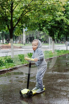 Drenched in the rain, a boy in a sport suit skates on a scooter. Spring walk in the city park, rainy weather.