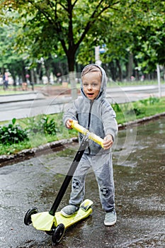 Drenched in the rain, a boy in a sport suit skates on a scooter. Spring walk in the city park, rainy weather.