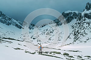 Oblečený muž s batohom v žiarivo oranžovej bunde s trekingovými palicami sa teší na výhľad na horské údolie počas horskej prechádzky vo vysokej