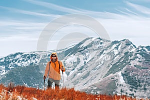 Dressed bright orange jacket backpacker walking by red blueberry field using trekking poles with mountain range background,