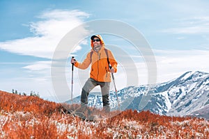 Oblečená zářivě oranžová bunda batohem chodící po červeném borůvkovém poli pomocí trekových holí s pozadím pohoří,
