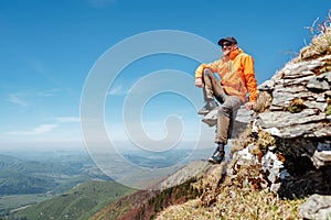 Dressed bright orange jacket hiker in baseball cap and sunglasses sitting on rocky cliff enjoying green valley at Mala Fatra