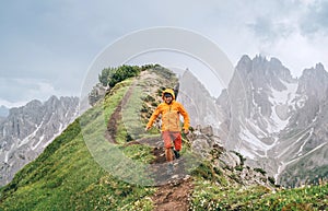 Dressed bright orange soft shell jacket backpacker running  by green mountain path with picturesque Dolomite Alps range background