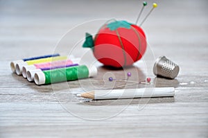 A dressmaker pencil with an out of focus pin cushion shaped as a red tomato, rolls of thread and a thimble