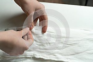 The dressmaker's hands sew white cotton fabric with a needle and thread on the work table. Close-up. Sewing Studio