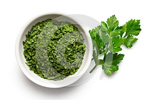 Dried chopped parsley in white ceramic bowl next to fresh parsley leaves isolated on white from above