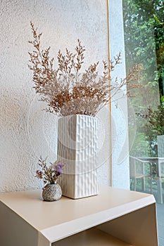 The dried flowers are housed in a white square vase, just set up next to a round iron item on a light wooden table on the edge of