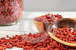 Dried goji berries in wooden bowls, scattered over white boards table under, blurred large glass jar full of fruits in background