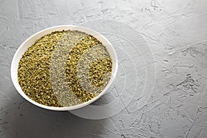 Dried Green Greek Oregano Spice in a white bowl on a gray surface, low angle view. Copy space