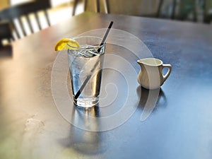 Drinking glass of cold ice water with lemon wedge and ceramic creamer on table