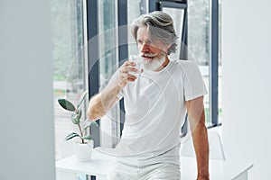 Drinks water. Senior stylish modern man with grey hair and beard indoors