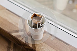 Drip bag in a paper cup on windowsill. Instant freshly brewed alternative drink in mug. Easy brew black coffee method