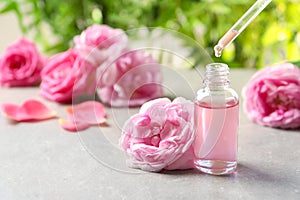 Dripping rose essential oil into bottle with pipette and fresh flowers on grey table