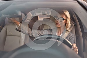 Drive safely. Beautiful business woman in eyeglasses sitting on the front seat of the car and adjusting rear-view mirror