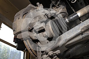 Drive shaft covered with rubber, visible disc and brake caliper, seen from the bottom of the car on a jack in the workshop.