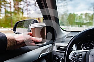 Driver holding paper cup with coffee at the car wheel.