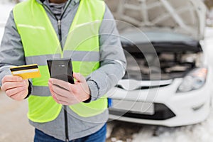 Driver holding road assistance service member card and smart phone. Broken car with opened hood in the background