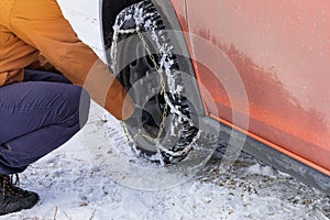 Driver installs chains on the wheel of his orange car in winter. Vehicle equipment for off-road.