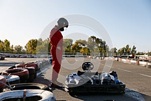 Driver wearing racing overalls and helmet standing nearby kart race car