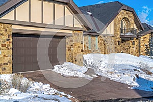 Driveway and snowy yard in front of garage door of home with stone brick wall