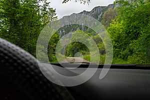 Driving car in the mountain in a path with green forest and big rocks. Driver view with black steering wheel