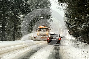 Driving on forest road through snowstorm, orange maintenance plough truck coming opposite way, view from car behind