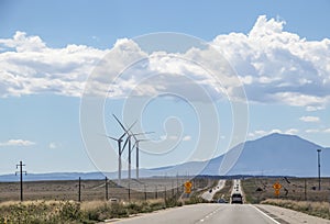 Driving on a long straight road with heat shimmer toward mountains - wind turbines on one side and signs that say Gusty Winds Area