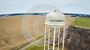 Drone aerial of empty field, partial forest in winter and green grass with Huntertown city water tower in Midwest