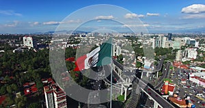 Drone-aerial view of a huge mexican flag waving. In the back, panoramic view of Mexico City. Many cars transit for the avenue.