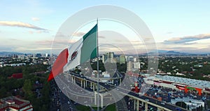 Drone-Aerial view of a huge mexican flag waving. In the back, panoramic viw of Mexico City. Many cars transit for the avenue.
