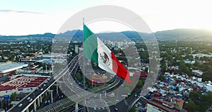 Drone-Aerial view of a huge mexican flag waving, at back the sun is hiding behind the mountains. Many cars transit for the avenue.