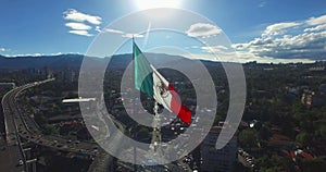 Drone-Aerial view of a huge mexican flag waving, the sun is up the flag. In the back, panoramic view of some suburbs and mountains