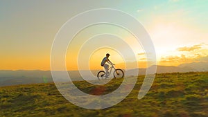 DRONE: Athletic tourist rides an electric bicycle up a grassy hill at sunrise