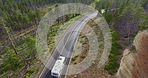 Drone camera takes off from a height a traveling white house on wheels and an oncoming black car on a highway in a pine forest