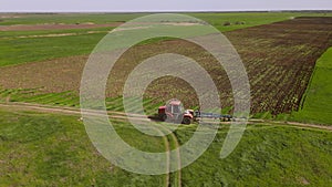 The drone flies around a red tractor with a plow, which drives along a dirt road along plowed agricultural field on one